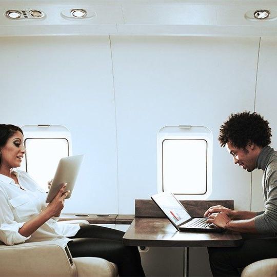 一男一女坐在私人飞机上的一张桌子旁，用笔记本电脑和平板电脑工作，并连接商务航空