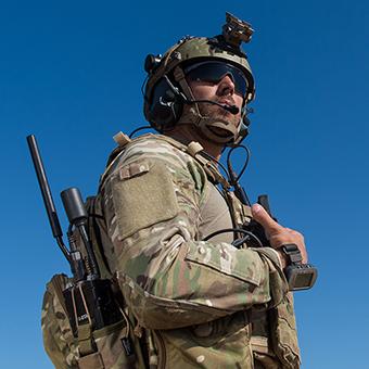 Soldier wearing green camo uniform, helmet, sunglasses and a backpack with a radio