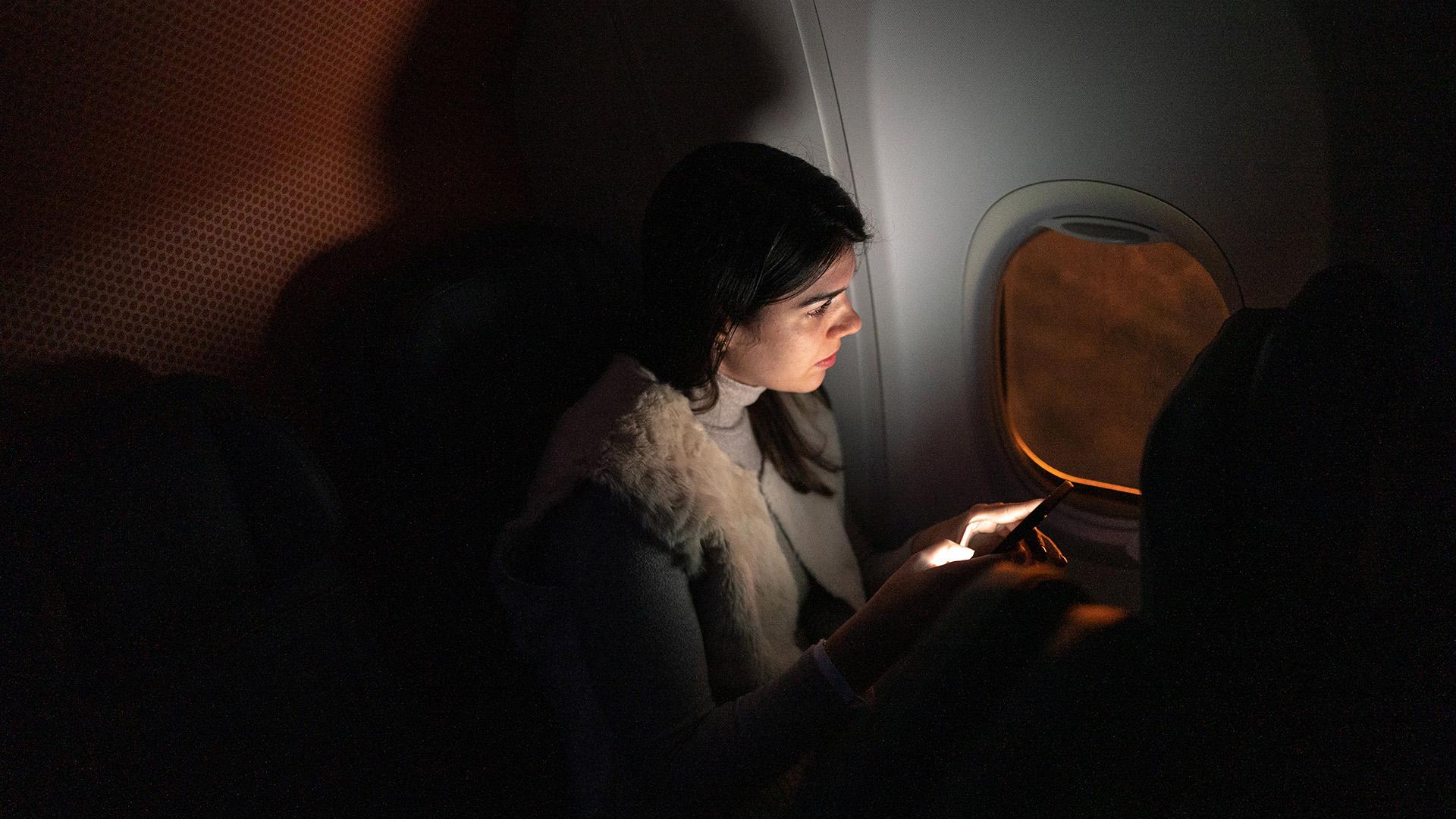 在昏暗的机舱里，一位留着长发的乘客坐在飞机窗户旁边, 眺望夕阳, 手机的光照亮了他们的脸.