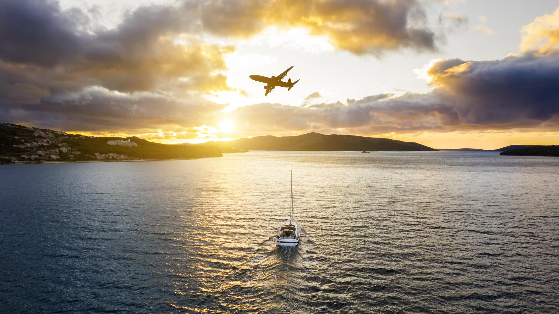 日落时分，一架客机从一艘帆船上方飞过，帆船独自在海上，靠近一些岛屿.