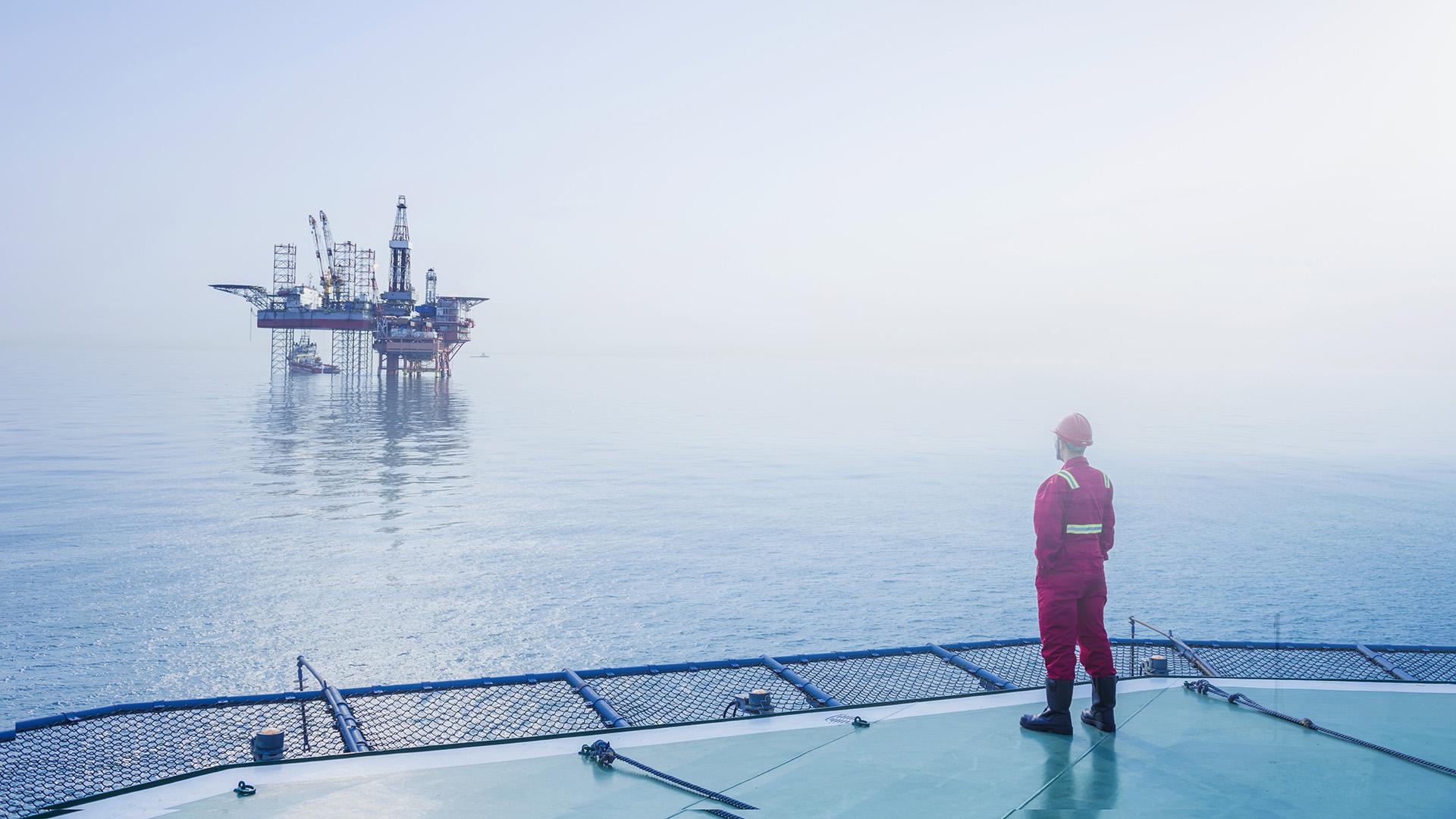 一个戴着安全帽、穿着工作服的人站在海上的一个平台上，望着对岸的石油钻井平台.